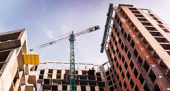 Vue du bas vers le haut d'un bâtiment en construction, avec une grue imposante opérant au-dessus, des ouvriers visibles en bas travaillant sur la façade et l'environnement autour du chantier.